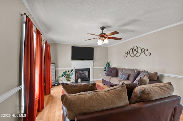 living room featuring light wood-style flooring, ceiling fan, a textured ceiling, a glass covered fireplace, and crown molding