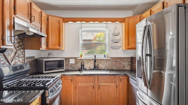 kitchen with a sink, dark stone countertops, under cabinet range hood, a textured ceiling, and appliances with stainless steel finishes