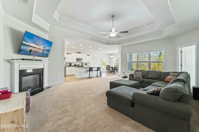 living room featuring ornamental molding, a ceiling fan, a tray ceiling, a glass covered fireplace, and light colored carpet