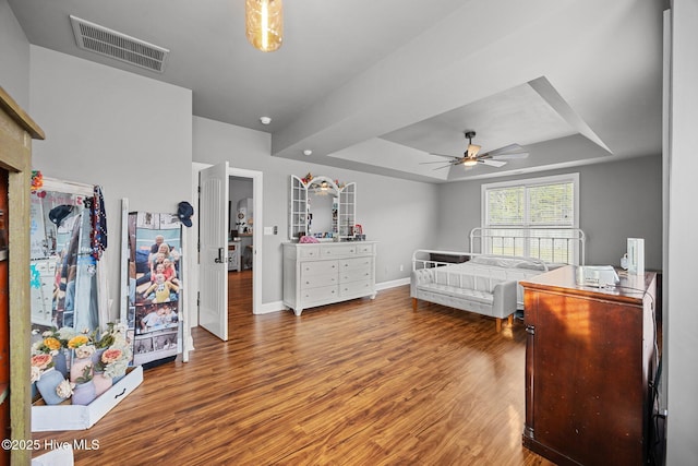 bedroom featuring wood finished floors, visible vents, baseboards, a tray ceiling, and ceiling fan