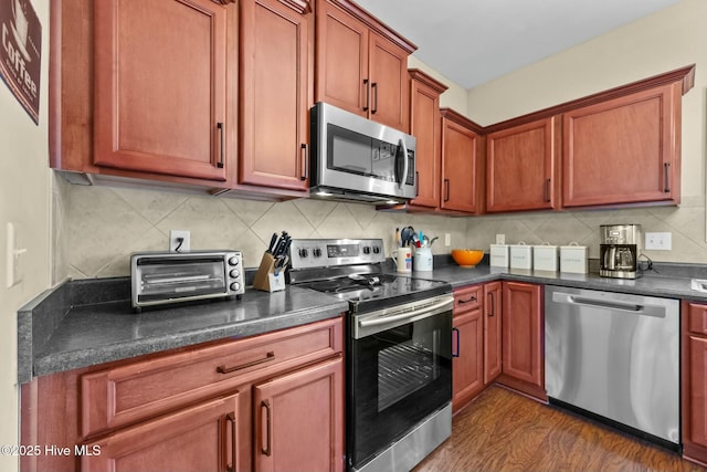 kitchen featuring dark wood-type flooring, backsplash, dark countertops, appliances with stainless steel finishes, and a toaster