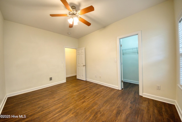 unfurnished bedroom featuring a walk in closet, baseboards, a ceiling fan, and dark wood-style flooring