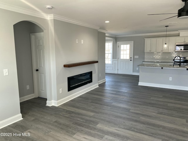 unfurnished living room with ornamental molding, a ceiling fan, a glass covered fireplace, dark wood finished floors, and arched walkways