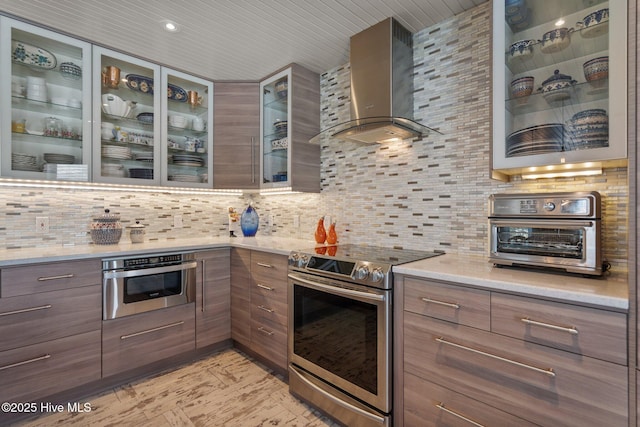 kitchen featuring backsplash, glass insert cabinets, appliances with stainless steel finishes, modern cabinets, and wall chimney exhaust hood