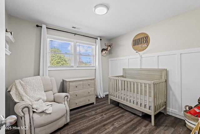 bedroom featuring dark wood-style floors, a wainscoted wall, visible vents, a nursery area, and a decorative wall