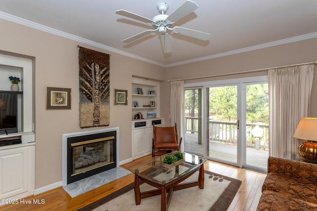 living area featuring light wood finished floors, ceiling fan, built in features, a fireplace with flush hearth, and ornamental molding