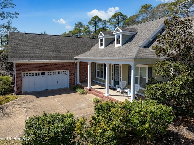 cape cod home with driveway, covered porch, an attached garage, a shingled roof, and brick siding