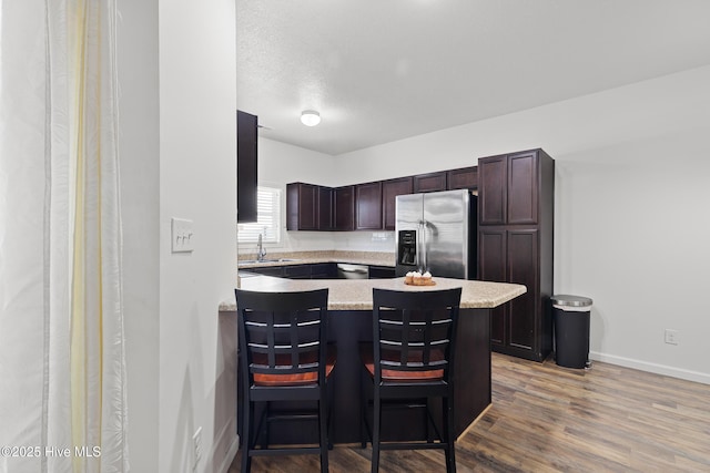 kitchen featuring dark brown cabinets, a peninsula, wood finished floors, stainless steel fridge, and a sink