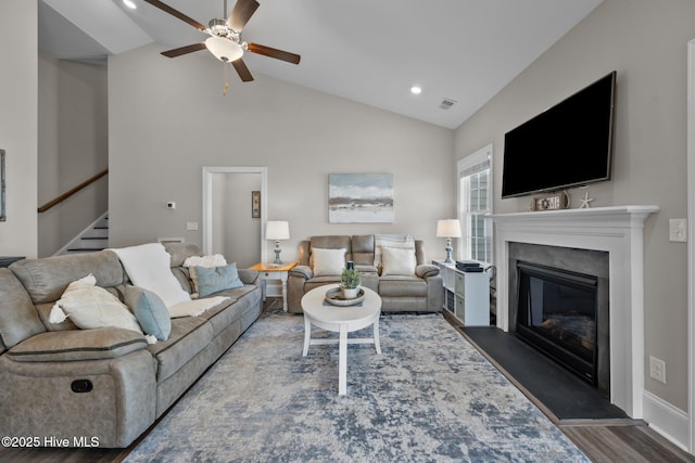 living area featuring visible vents, stairway, vaulted ceiling, a glass covered fireplace, and a ceiling fan