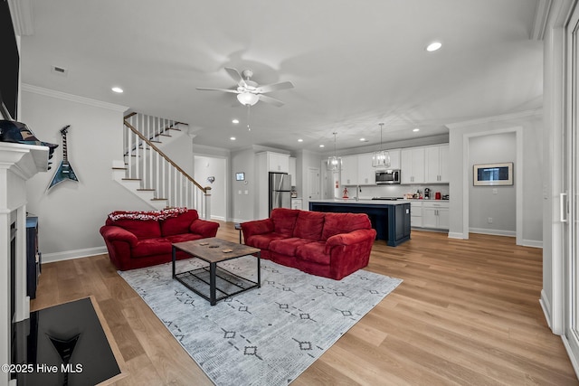 living area featuring stairway, light wood-type flooring, ornamental molding, ceiling fan with notable chandelier, and a fireplace