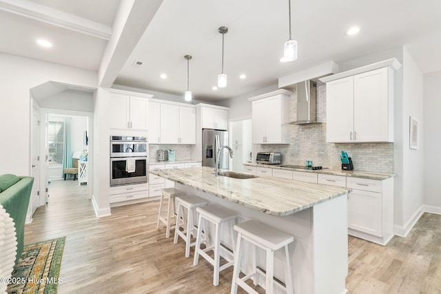 kitchen featuring a sink, light wood-style floors, appliances with stainless steel finishes, a kitchen bar, and wall chimney range hood