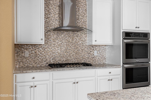 kitchen featuring light stone counters, stainless steel appliances, decorative backsplash, white cabinetry, and wall chimney range hood