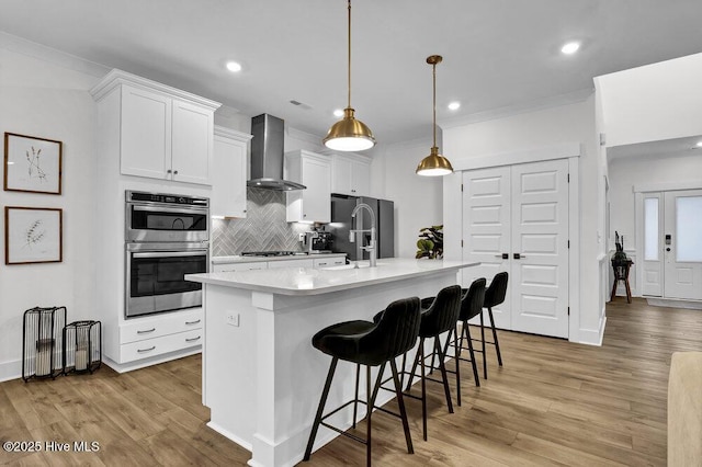 kitchen featuring backsplash, wall chimney range hood, light countertops, light wood-style flooring, and stainless steel appliances