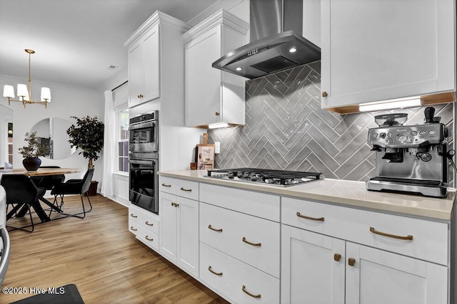 kitchen with light wood-type flooring, light countertops, white cabinets, wall chimney range hood, and tasteful backsplash