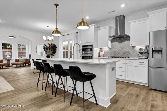 kitchen featuring light wood-style flooring, light countertops, appliances with stainless steel finishes, wall chimney exhaust hood, and tasteful backsplash