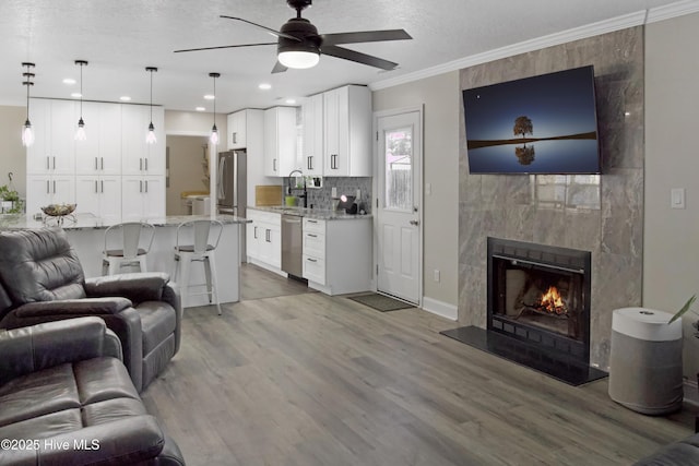 living room with light wood-type flooring, a tiled fireplace, a textured ceiling, crown molding, and ceiling fan