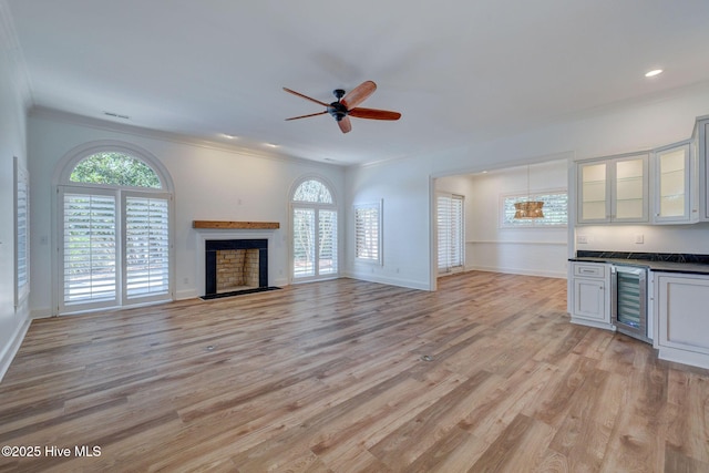 unfurnished living room with light wood-type flooring, beverage cooler, a fireplace, crown molding, and ceiling fan