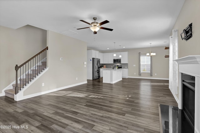 unfurnished living room with dark wood-type flooring, baseboards, stairway, ceiling fan with notable chandelier, and a fireplace