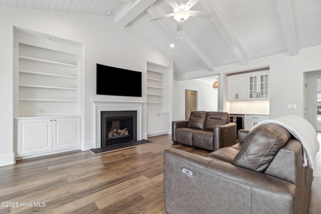 living area with lofted ceiling with beams, a ceiling fan, a glass covered fireplace, wood finished floors, and built in shelves