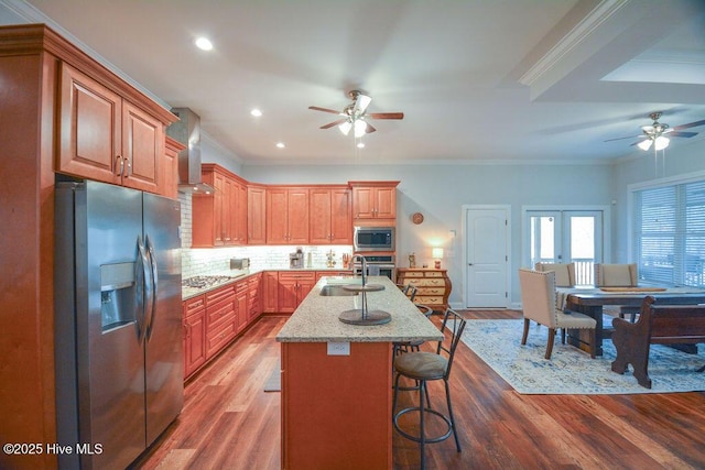 kitchen featuring a center island with sink, stainless steel appliances, wall chimney exhaust hood, and ceiling fan