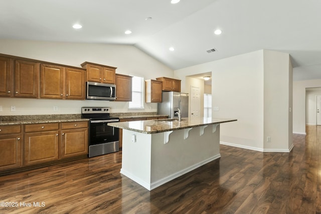 kitchen with dark stone counters, a kitchen island with sink, appliances with stainless steel finishes, a kitchen bar, and brown cabinets