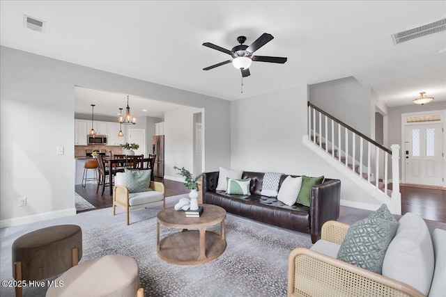 living room with stairway, baseboards, visible vents, dark wood finished floors, and ceiling fan with notable chandelier