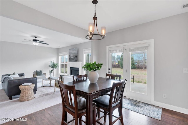 dining area featuring dark wood finished floors, ceiling fan with notable chandelier, a fireplace, and baseboards