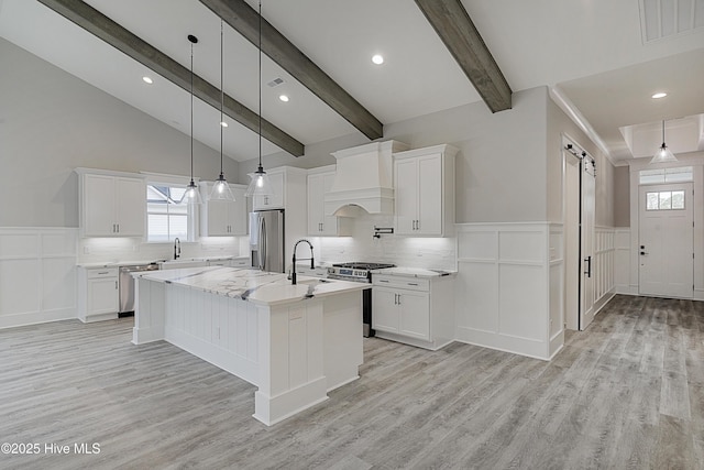 kitchen featuring appliances with stainless steel finishes, light wood-style floors, custom exhaust hood, white cabinets, and a kitchen island with sink