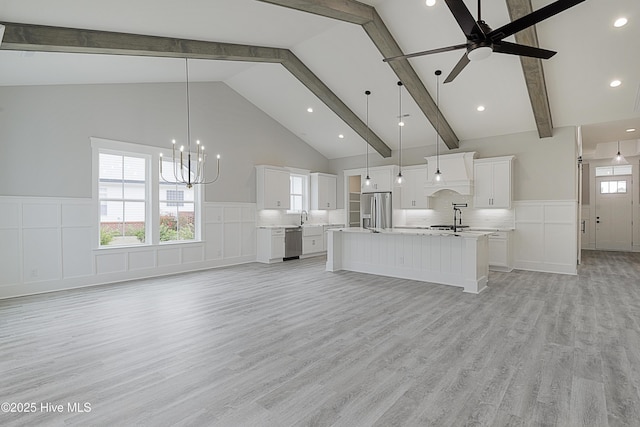 unfurnished living room with beam ceiling, high vaulted ceiling, light wood-style flooring, ceiling fan with notable chandelier, and wainscoting