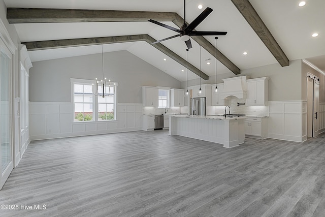 unfurnished living room featuring light wood finished floors, a wainscoted wall, beam ceiling, and ceiling fan with notable chandelier