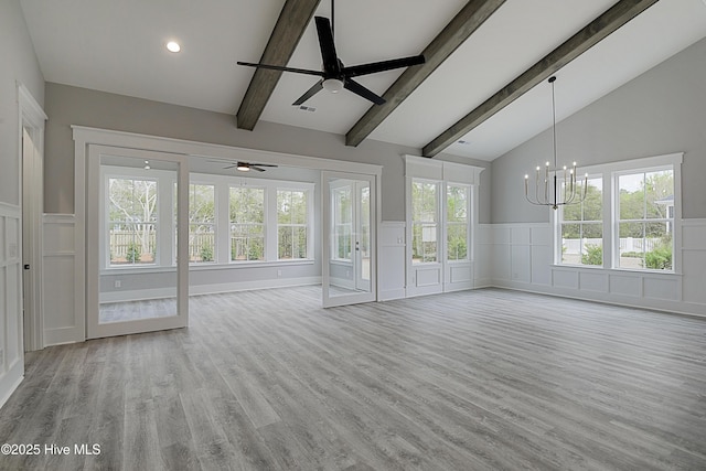 unfurnished living room with ceiling fan with notable chandelier, wainscoting, vaulted ceiling with beams, and light wood finished floors