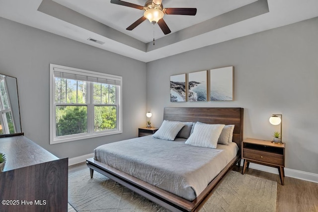 bedroom with visible vents, wood finished floors, baseboards, ceiling fan, and a tray ceiling