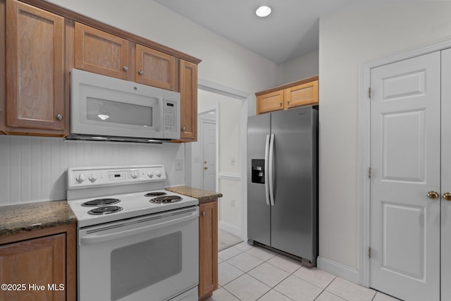 kitchen with brown cabinetry, light tile patterned floors, baseboards, dark stone countertops, and white appliances