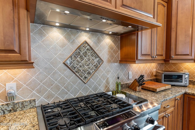 kitchen with brown cabinets, backsplash, stainless steel range with gas cooktop, light stone countertops, and extractor fan