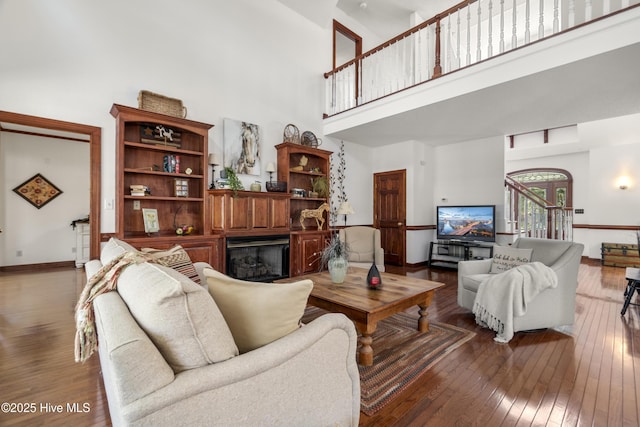 living room featuring wood-type flooring, a towering ceiling, a glass covered fireplace, and baseboards