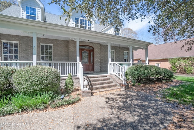 doorway to property featuring covered porch, brick siding, and roof with shingles