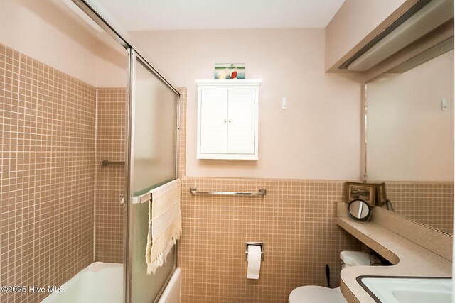 full bathroom featuring toilet, bath / shower combo with glass door, a wainscoted wall, and tile walls