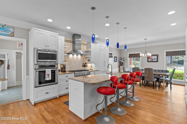 kitchen with light wood-style floors, wall chimney exhaust hood, ornamental molding, appliances with stainless steel finishes, and a sink