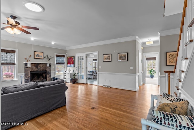 living room featuring a stone fireplace, ceiling fan, crown molding, stairs, and light wood-style flooring