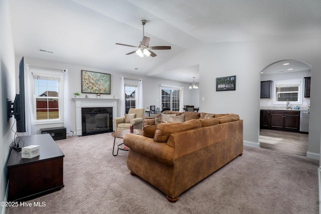 living room featuring vaulted ceiling, a glass covered fireplace, light colored carpet, arched walkways, and ceiling fan