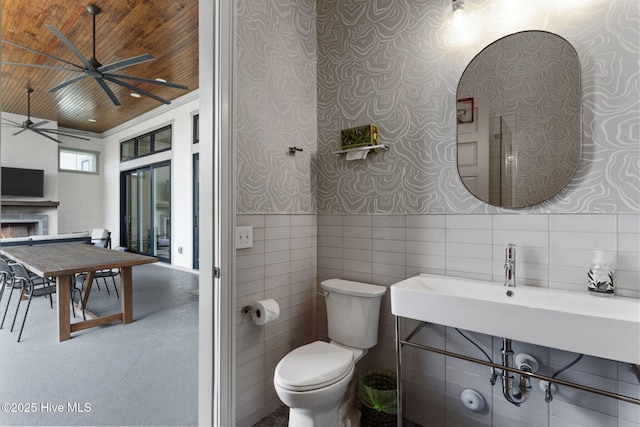bathroom featuring a lit fireplace, wallpapered walls, toilet, ceiling fan, and wood ceiling