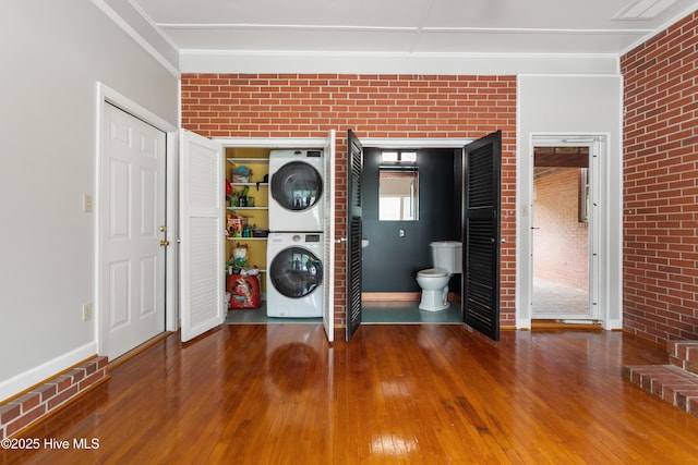 clothes washing area featuring laundry area, brick wall, hardwood / wood-style flooring, baseboards, and stacked washing maching and dryer