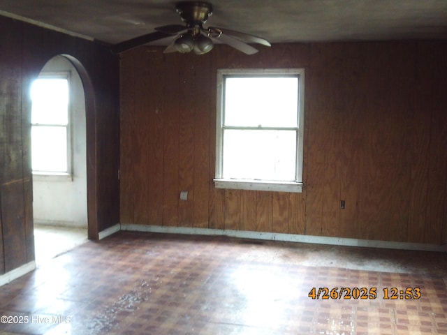 unfurnished room featuring wooden walls, arched walkways, a ceiling fan, and a wealth of natural light