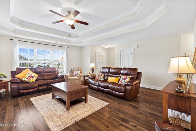 living room featuring dark wood-style flooring, baseboards, ceiling fan, ornamental molding, and a tray ceiling