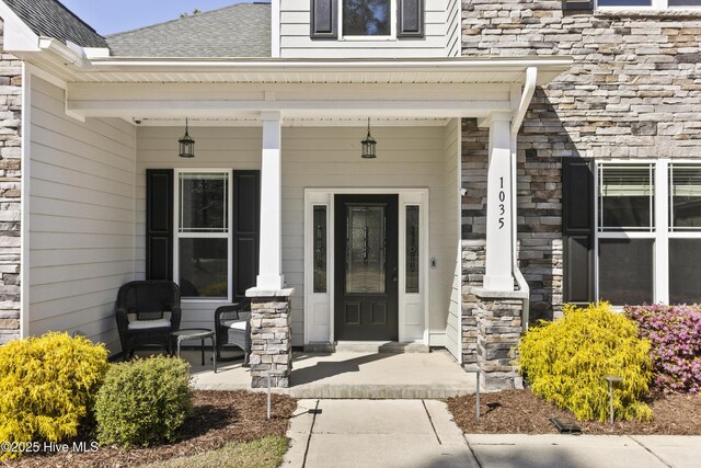 doorway to property featuring a shingled roof, a porch, and stone siding