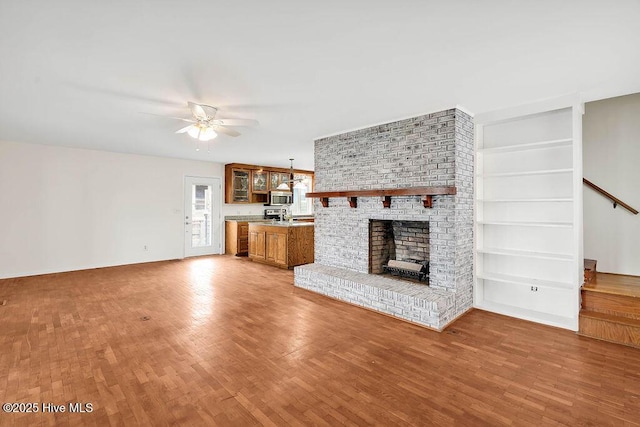 unfurnished living room featuring built in shelves, a brick fireplace, ceiling fan, light wood finished floors, and stairs