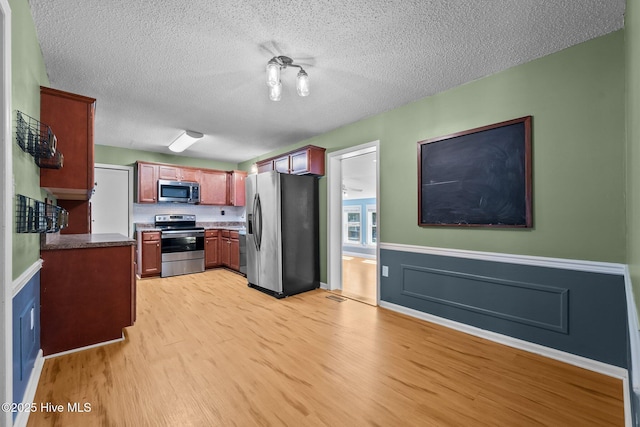 kitchen with a wainscoted wall, a textured ceiling, stainless steel appliances, and light wood-style floors