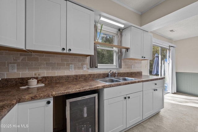 kitchen with white cabinets, dark countertops, a sink, beverage cooler, and wainscoting