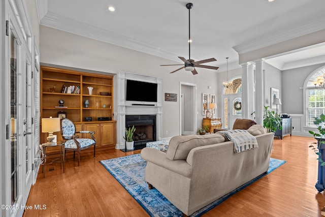 living room featuring crown molding, light wood finished floors, a fireplace, ceiling fan with notable chandelier, and decorative columns