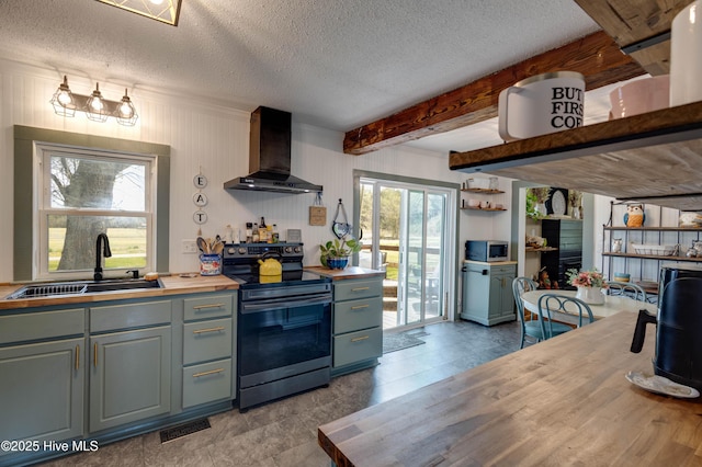 kitchen featuring wooden counters, wall chimney exhaust hood, beam ceiling, appliances with stainless steel finishes, and a sink